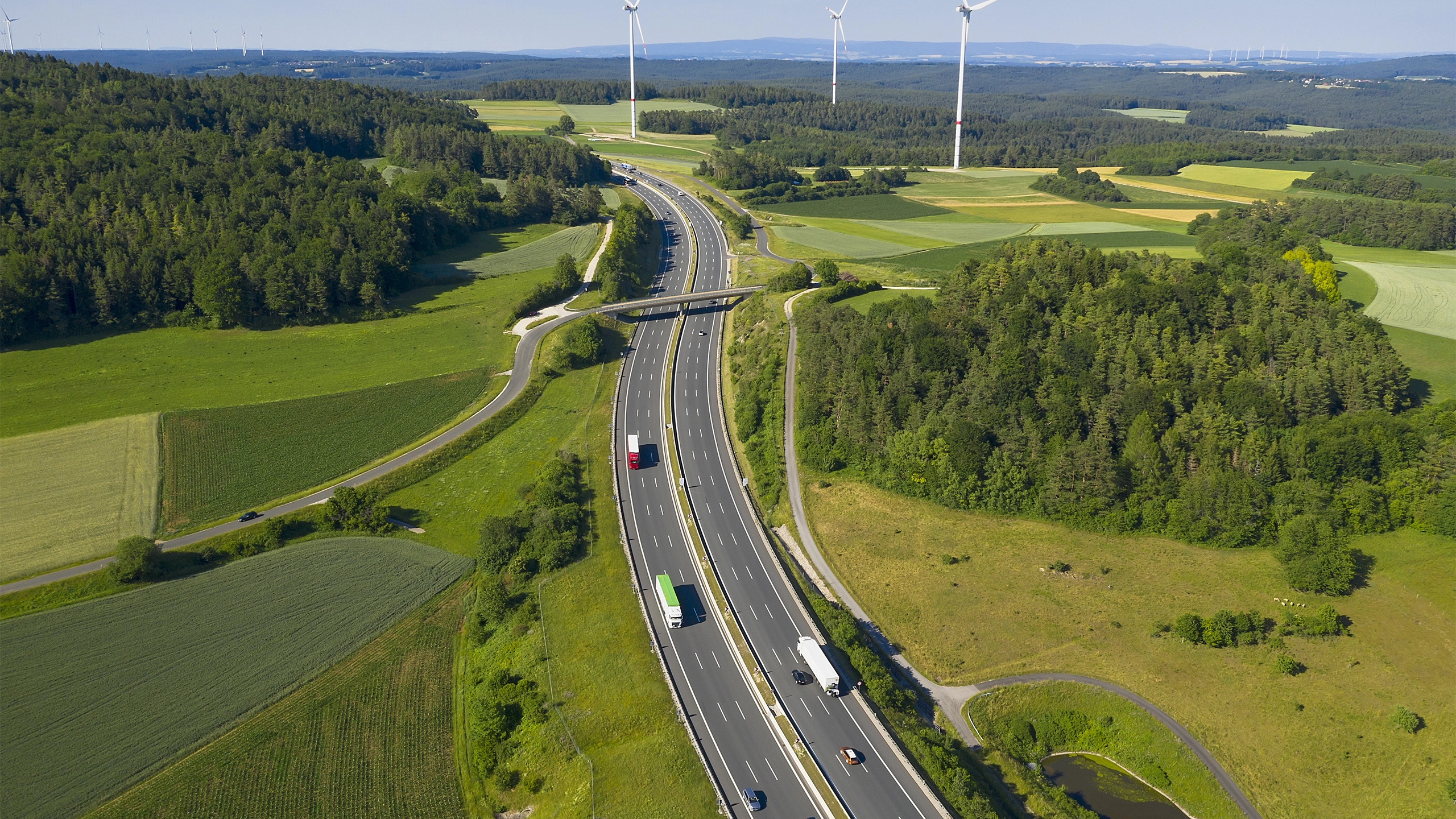 Traffic on a motorway surrounded by green fields, forest and wind turbines in the far distance