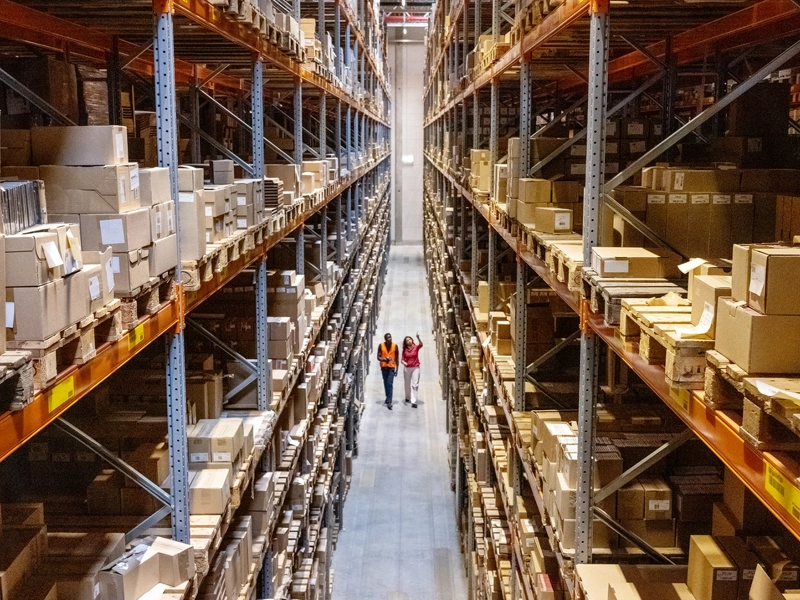 Two employees in high vis jackets walking through warehouse with tall shelves full of products
